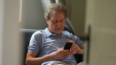 Telefon kullanan dürüst yaşlı bir adam, koltukta oturmuş akıllı telefona bakan yaşlı bir adam.