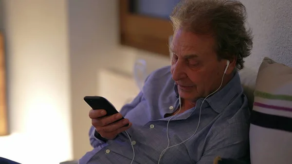 Senior man talking on phone wearing earphones at home. Older person talks on cellphone