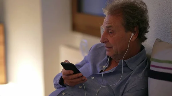 Senior man talking on phone wearing earphones at home. Older person talks on cellphone
