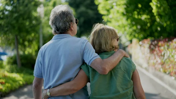 Romantic senior husband putting arm around wife, back view of older couple walking together in green path