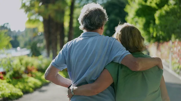 Romantic senior husband putting arm around wife, back view of older couple walking together in green path