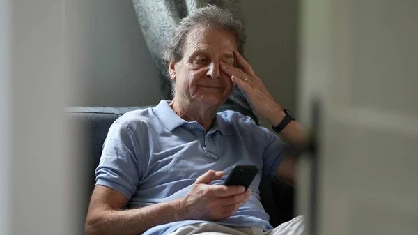 Pensive senior man sitting on sofa looking at cellphone device, person scratching eyes