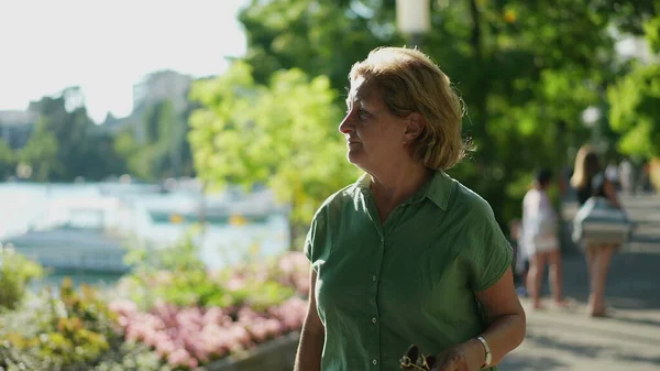 Older woman walking outdoors in green urban city, senior person walks forward outside