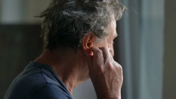 Older man touching ear scratching earlobe standing by window