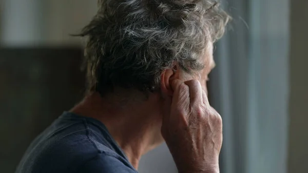 Older man touching ear scratching earlobe standing by window