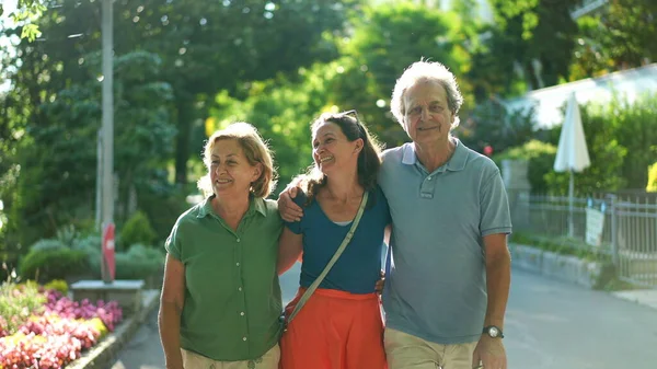 Joyful daughter walking with senior parents together in green city laughing and smiling