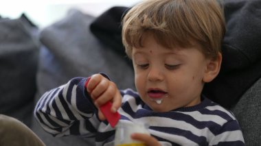 Kanepede oturan yoğurt yiyen bir çocuk kaşıkla yoğurt yiyor.