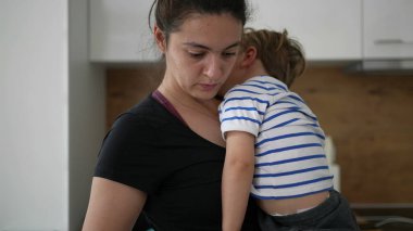 Anne oğlunu kucağında tutarken anne evde yemek hazırlıyor.