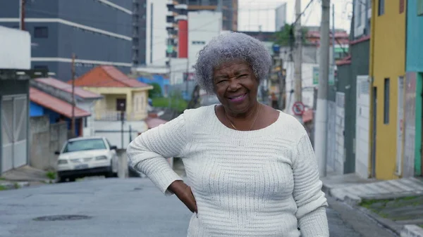 An older black woman standing outside in urban street looking at camera