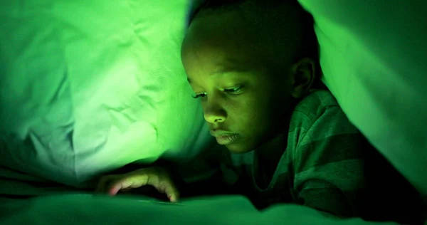 Young boy looking at cellphone screen at night under blanket
