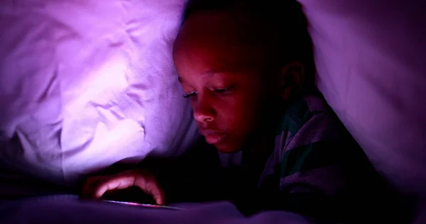 Young boy looking at cellphone screen at night under blanket