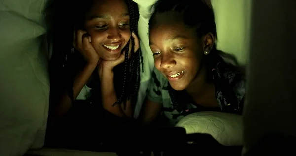 Two African teen girls looking at smartphone screen at night under blanket, real life laugh and smile reaction to video content