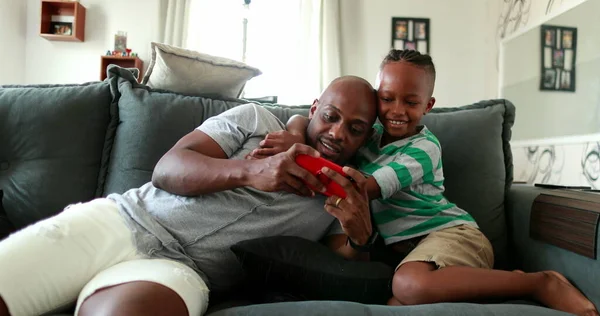 Black father and child together looking at cellphone video. African mixed race parent and little boy hug embrace at sofa