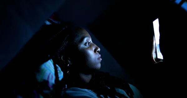 Black African teen girl lying on bed looking at cellphone screen at night