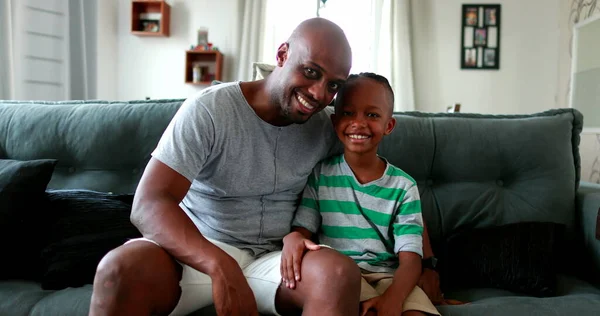 African father and son sitting on couch smiling at camera
