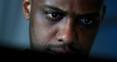 Tired black man working late at night in front of computer. Close-up face looking at screen focused and concentrated