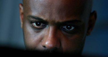 Tired black man working late at night in front of computer. Close-up face looking at screen focused and concentrated