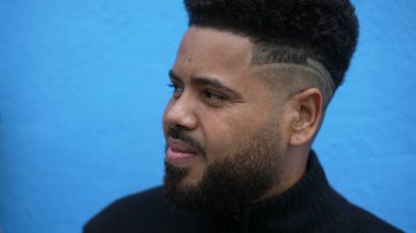 Mavi arka planda, modern saç kesimiyle sokak gözlemcisi dalgın bir genç.