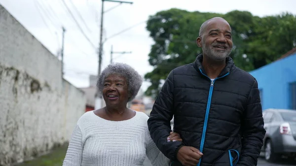 An aged son walking with elderly mother in 80s outside in urban street going for a walk