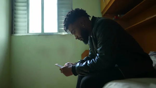 A young black man staring at phone screen sitting in bedroom using smartphone