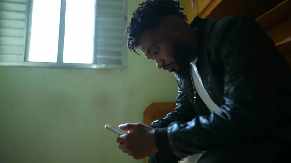 A young black man staring at phone screen sitting in bedroom using smartphone