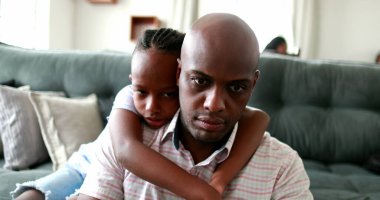 Evde çalışan siyah bir baba, bilgisayarın önünde ebeveyni kucaklayan bir çocuk.