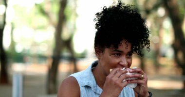 Siyah bir kadın parkta hamburger yiyor.