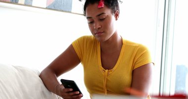 Evdeki koltukta akıllı telefon kullanan siyah bir kadın.