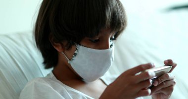 Çocuk internette oyun oynuyor. Salgın sırasında akıllı telefon tutuyor. Evde maske takıyor.