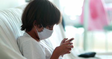 Çocuk salgın sırasında cep telefonuyla video oyunu oynuyor, covid-19 maskesi takıyor.