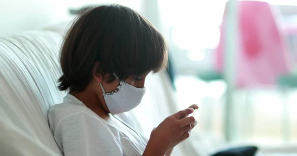 Boy playing video-game on cellphone during pandemic, wearing covid-19 mask