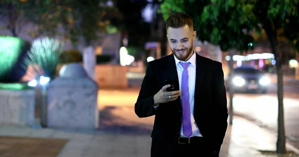 Handsome business man checking smartphone at night smiling. Corporate executive walking in city