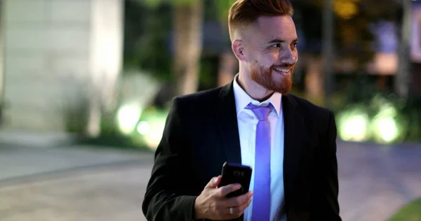 Handsome business man checking smartphone at night smiling. Corporate executive walking in city