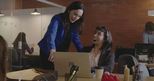 Executive female leader chief helping younger employer wearing headset in front of laptop computer at corporate modern office