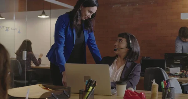 Executive female leader chief helping younger employer wearing headset in front of laptop computer at corporate modern office