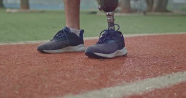 Closeup of person with prosthetic leg and tennis shoes standing at running track with equipment