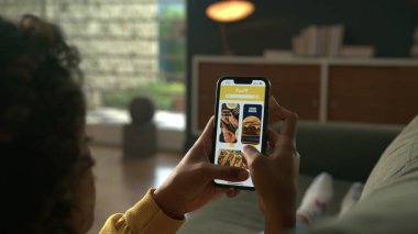 Çevrimiçi Dağıtım menü telefonunu kullanan kişi. Online mağazadaki kadın siparişleri teslimat uygulamasında akıllı telefon kullanıyor. Oturma odasındaki kanepede uzanan kadın.