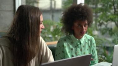Two young diverse female colleagues working at coffee shop in front of laptop computer