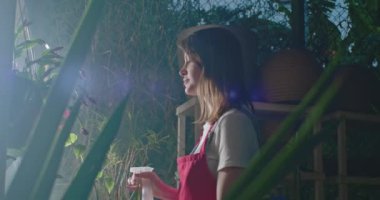 Young woman spraying water into plants inside greenhouse. Employee of small business caring for plants and flowers irrigating. Girl wearing apron watering plants