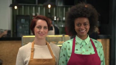 Two young diverse workers portrait standing inside coffee shop. Local business concept of female baristas wearing aprons smiling