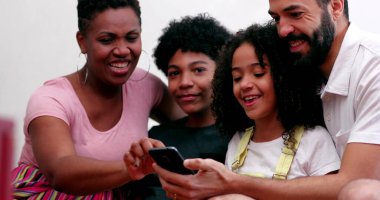 Irklar arası aile ve çocuklar birlikte gülüyor. Ev koltuğunda cep telefonuna bakıyorlar.