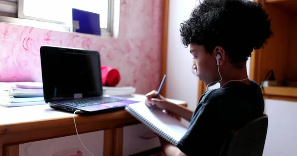 Teen boy studying at home in front of laptop computer. Mixed race kid writing notes on notepad