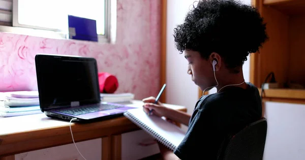 Teen boy studying at home in front of laptop computer. Mixed race kid writing notes on notepad