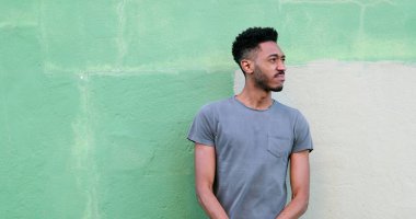 Genç siyahi adam dışarıda kollarını kavuşturup duvara yaslanıyor ve rastgele kameraya bakıyor.