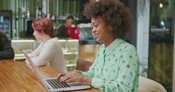 A happy black woman using laptop at coffee shop. One African American girl typing on computer keyboard smiling. A female entrepreneur person looking at laptop screen