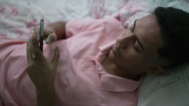 A young hispanic man looking at cellphone device in bedroom lying in bed