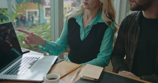 Senior executive female leader coaching younger employee in front of laptop. Closeup hand pointing at computer screen manager explaining job to younger staff at coffee shop