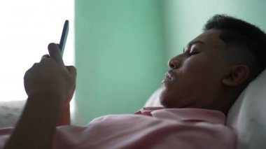 A Brazilian young man lying in bed texting message on smartphone device. One hispanic south american person using phone