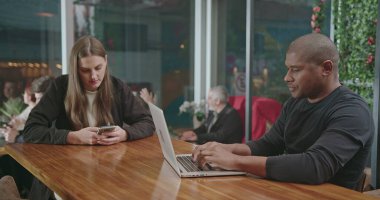 İnsanlar kafeteryada modern teknoloji cihazları kullanarak oturuyorlar. Bilgisayarda yazan dizüstü bilgisayarı açan siyah bir adam. Kafedeki akıllı telefon cihazına bakan kız