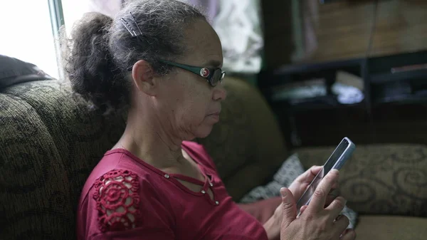 A south american older lady sitting at home using technology cellphone device
